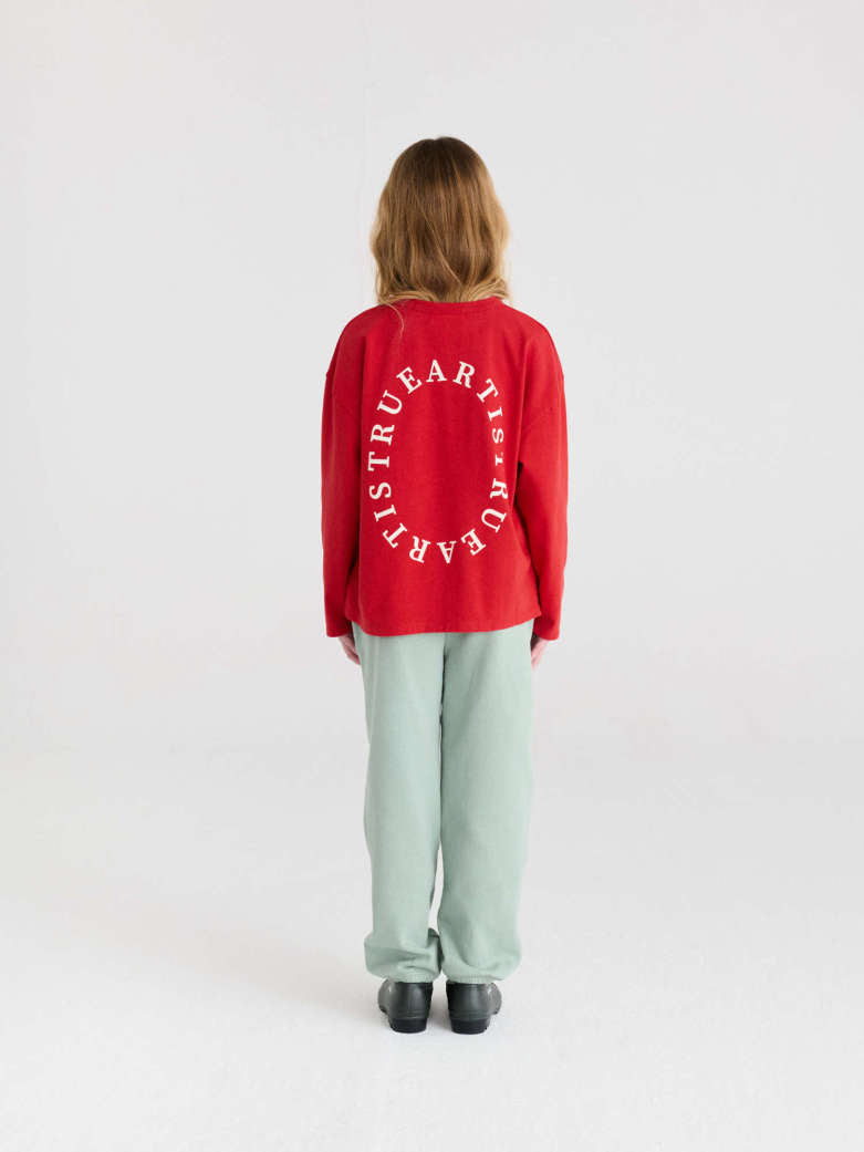 A model viewed from behind wearing a bright red sweatshirt with a white circular graphic text print on the back, paired with sage green sweatpants and black shoes, against a plain white studio background.