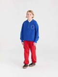 A child model wearing a royal blue quarter-zip sweatshirt with a white circular logo on the chest, paired with bright red sweatpants and brown shoes, standing against a seamless white studio background.