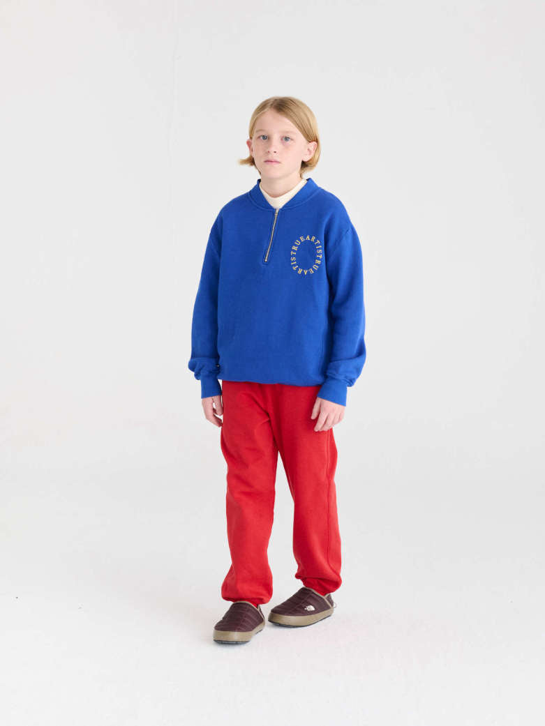 A child model wearing a royal blue quarter-zip sweatshirt with a white circular logo on the chest, paired with bright red sweatpants and brown shoes, standing against a seamless white studio background.