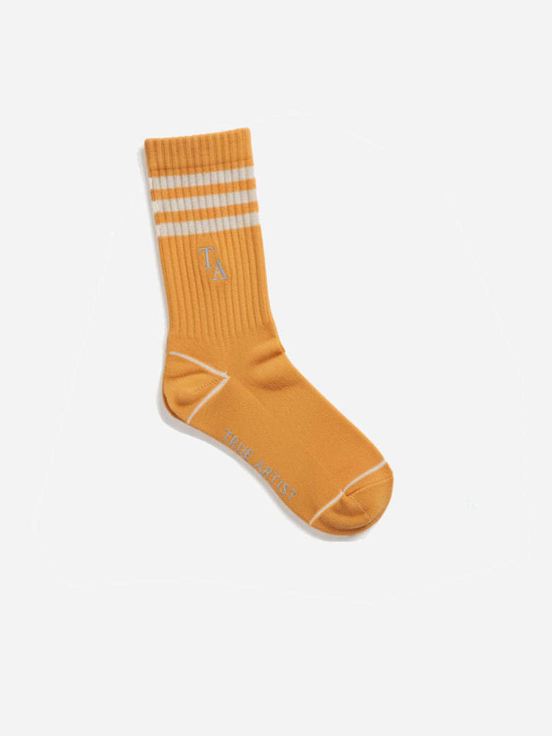 A single mustard yellow crew sock with three white stripes on the cuff and a small white embroidered logo, displayed on a clean white background.