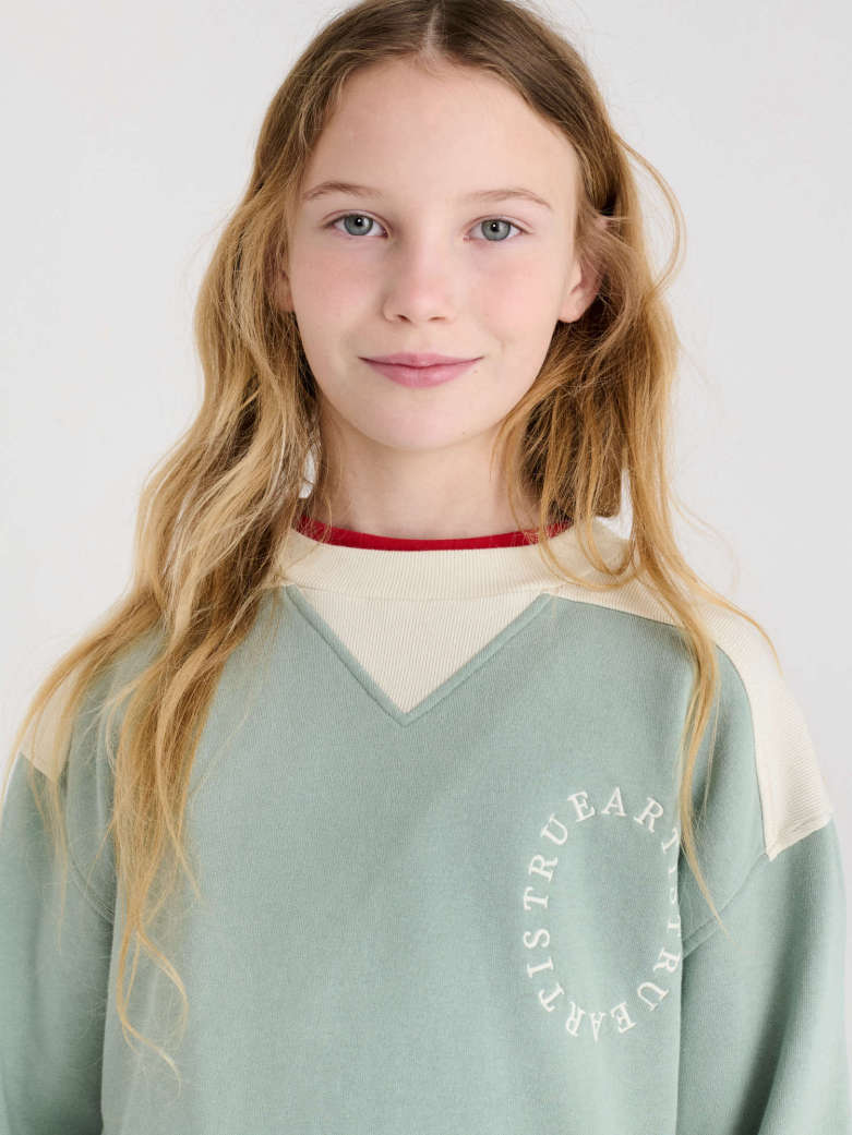 A young girl with long blonde hair wearing a sage green and cream colorblock sweatshirt with a red inner collar, shown against a plain light gray background.