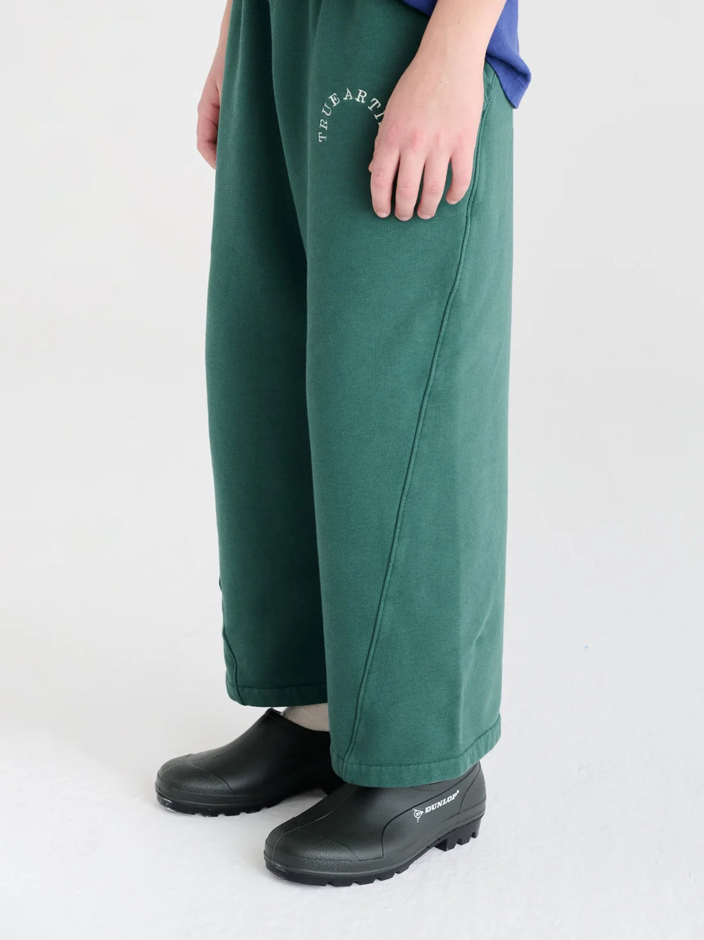 A person wearing wide-leg green pants with a small white circular logo on the upper left thigh, paired with black clogs, shown from the waist down against a light gray studio background.