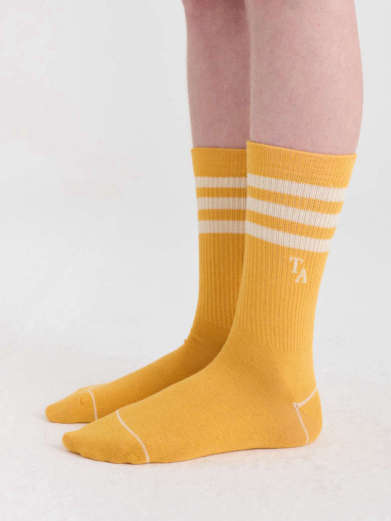 A pair of mustard yellow crew socks with white stripes and a small white embroidered logo, shown on a person's feet against a plain light background.