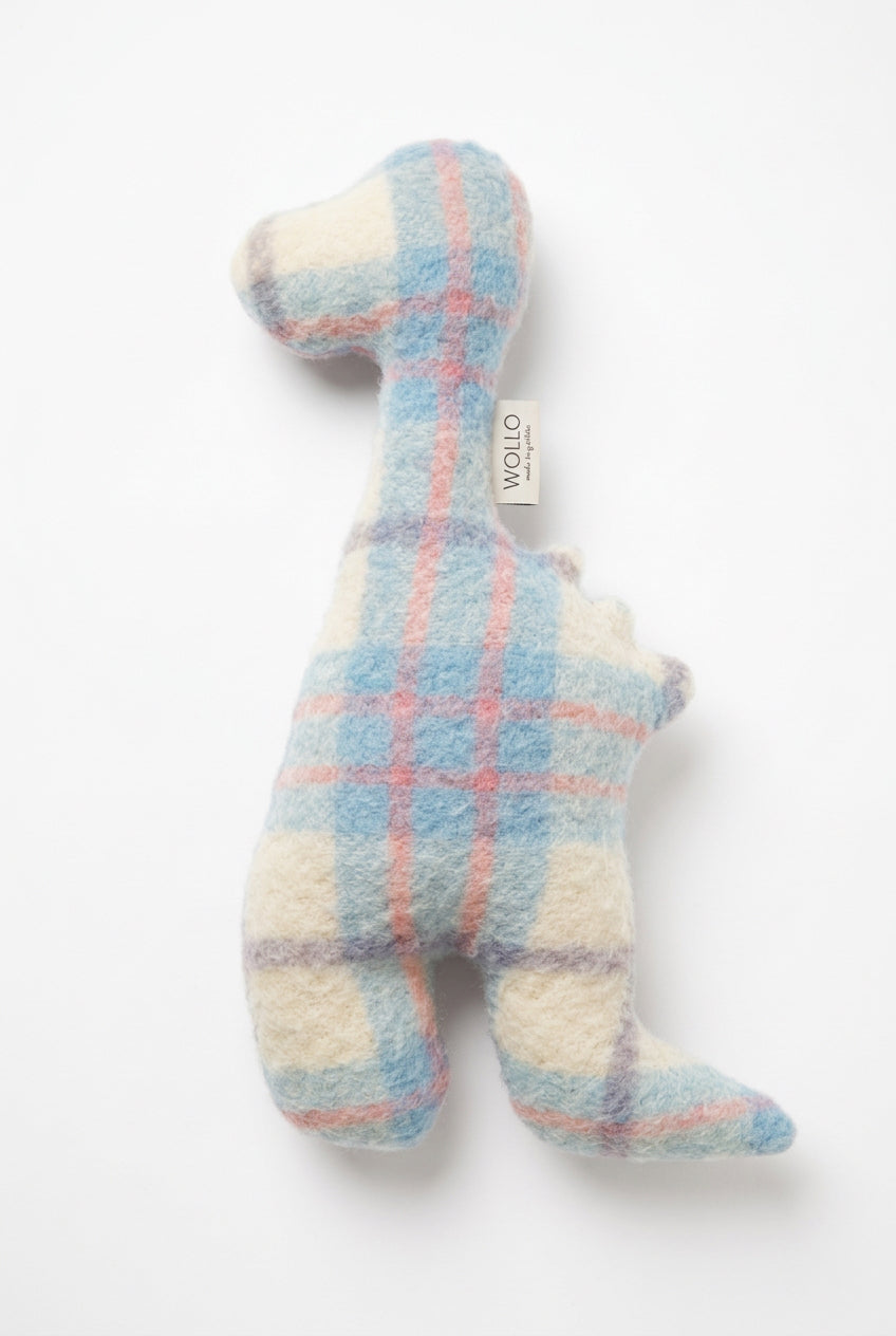 A handmade wool felt dinosaur plush toy in a pastel plaid pattern of light blue, cream, and pink, lying on a wooden floor. A small fabric tag with the brand name 'WOOLD' is visible near the neck.