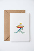 A white greeting card featuring a watercolor illustration of a small red boat with a yellow flag riding a green wave, placed on top of a brown kraft envelope on a wooden surface.