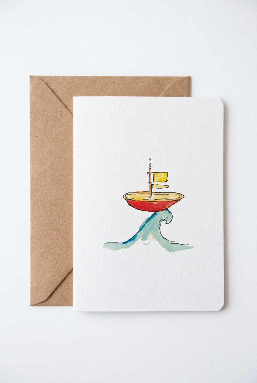 A white greeting card featuring a watercolor illustration of a small red boat with a yellow flag riding a green wave, placed on top of a brown kraft envelope on a wooden surface.