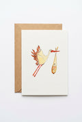 A white art print card featuring a whimsical watercolor illustration of a white stork carrying a baby bundle, shown with a brown kraft envelope on a clean white background.