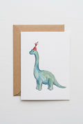 A watercolor dinosaur greeting card featuring a blue and green long-necked dinosaur with a red party hat, shown with a brown kraft envelope on a wooden table.