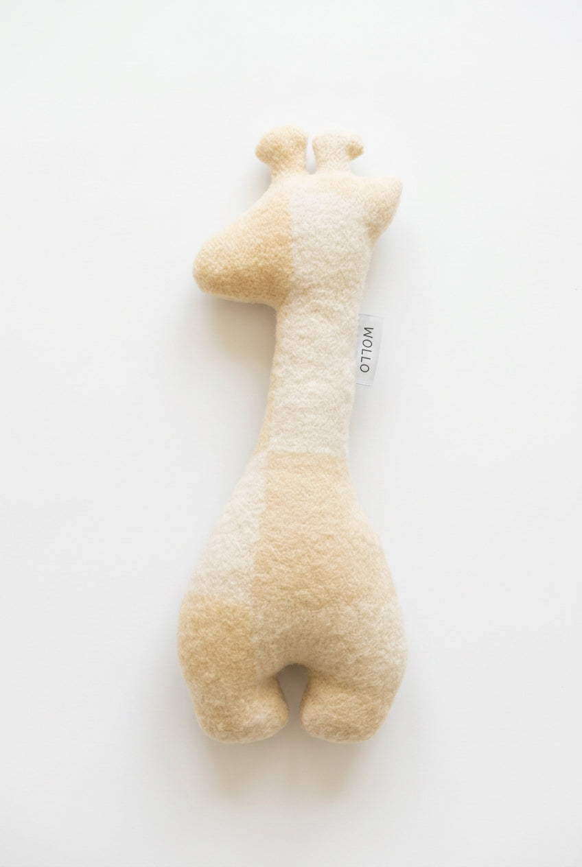 A plush giraffe toy in a cream and pale yellow colorway, lying flat on a light brown wooden floor. A small white tag with the brand name 'HOLA' is visible on the side.