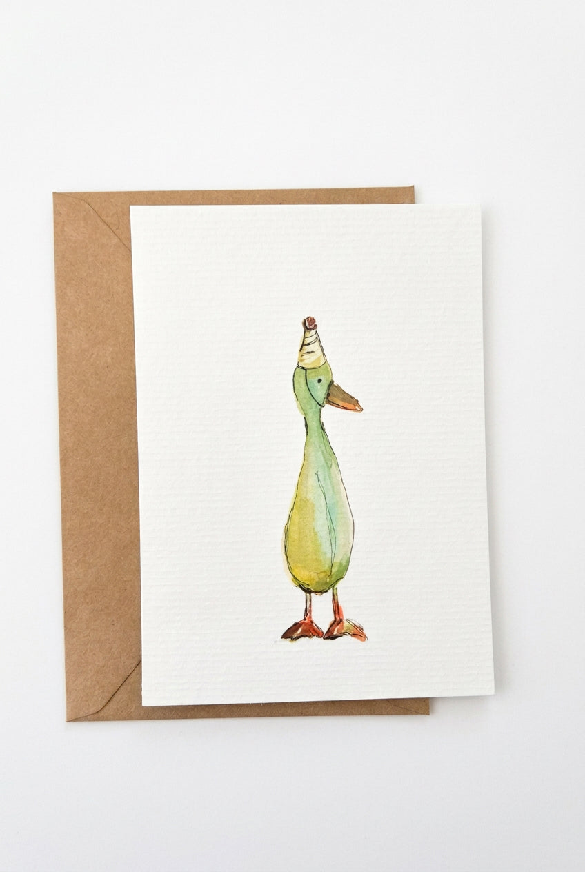A white greeting card featuring a watercolor illustration of a duck wearing a party hat, shown with a brown kraft envelope on a clean white background.