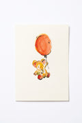 A white greeting card with a watercolor illustration of a teddy  elephant holding an orange balloon, placed on top of a brown kraft envelope on a wooden table.