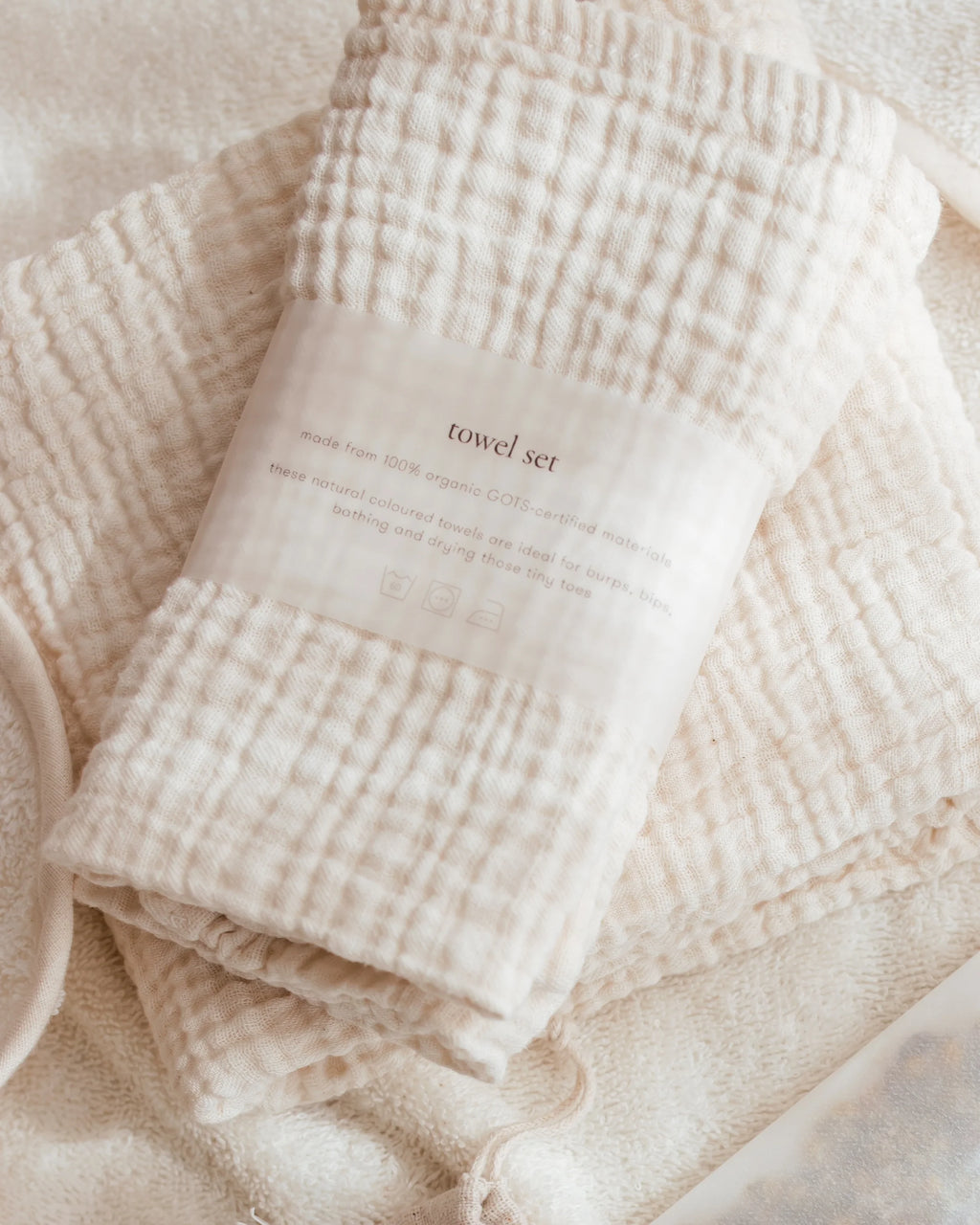 A folded cotton muslin towel set in a warm cream color, wrapped with a paper band label that reads 'towel set'. The towels are arranged on a soft, textured cream fabric background.