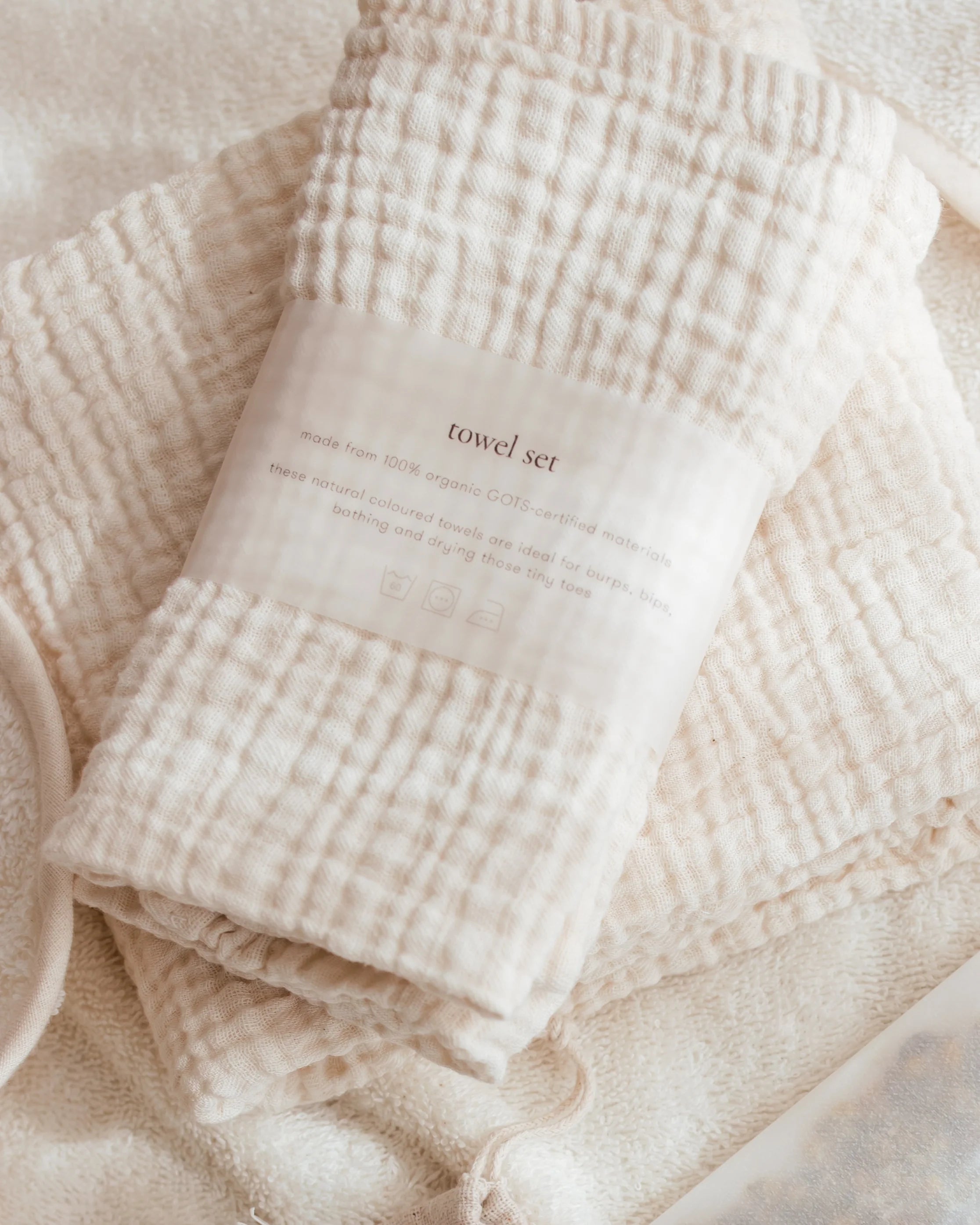 A folded cotton muslin towel set in a warm cream color, wrapped with a paper band label that reads 'towel set'. The towels are arranged on a soft, textured cream fabric background.