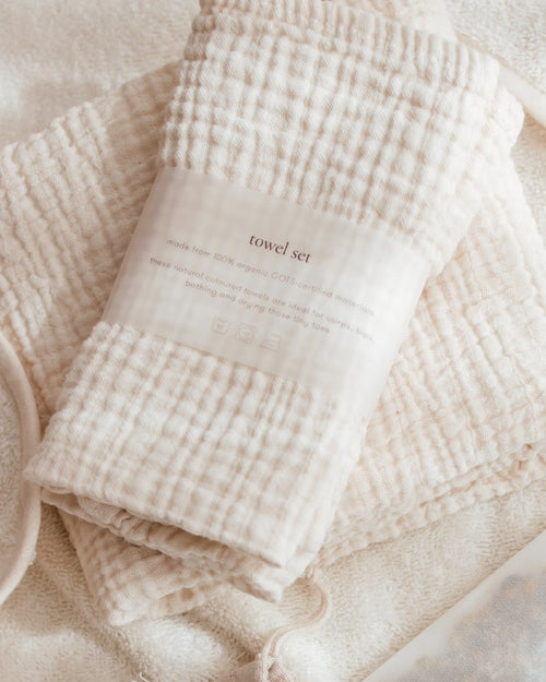 A folded cotton muslin towel set in a warm cream color, wrapped with a paper band label that reads 'towel set'. The towels are arranged on a soft, textured cream fabric background.