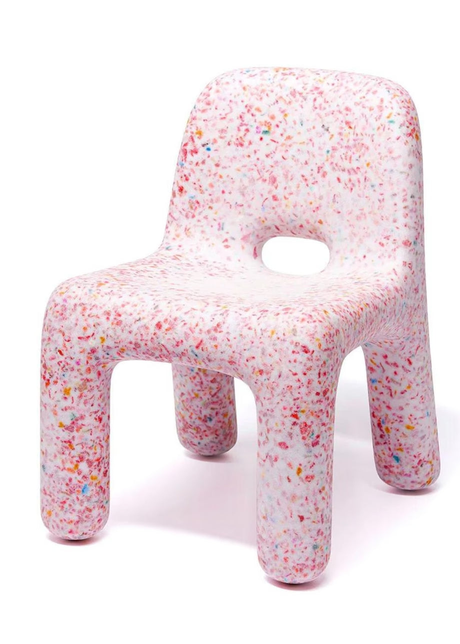 A modern molded recycled chair with a rounded, blocky silhouette, featuring a colorful confetti speckled pattern on a white base. The chair has a cutout in the center of the backrest and is photographed on a seamless white background.