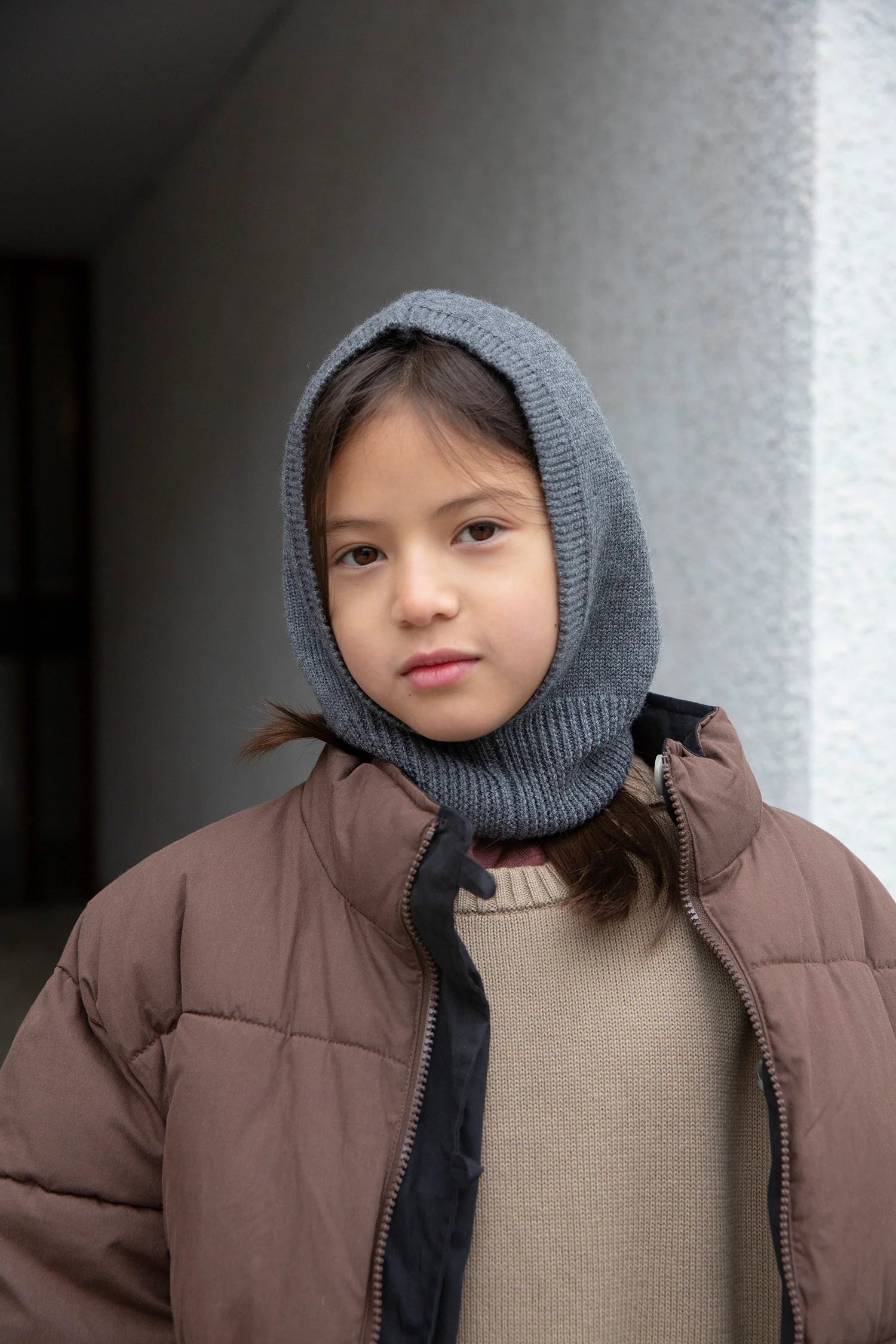 A child wearing a brown puffer jacket, a beige knit sweater, and a gray ribbed knit hooded neck warmer, standing against a textured gray wall.