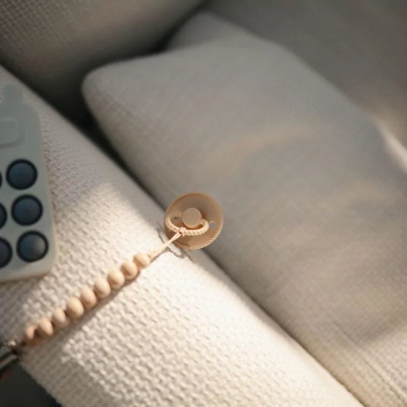 A close-up of a beige fabric accessory with a small round button and a beaded cord, resting on a textured cream fabric pacifier surface next to a remote control.