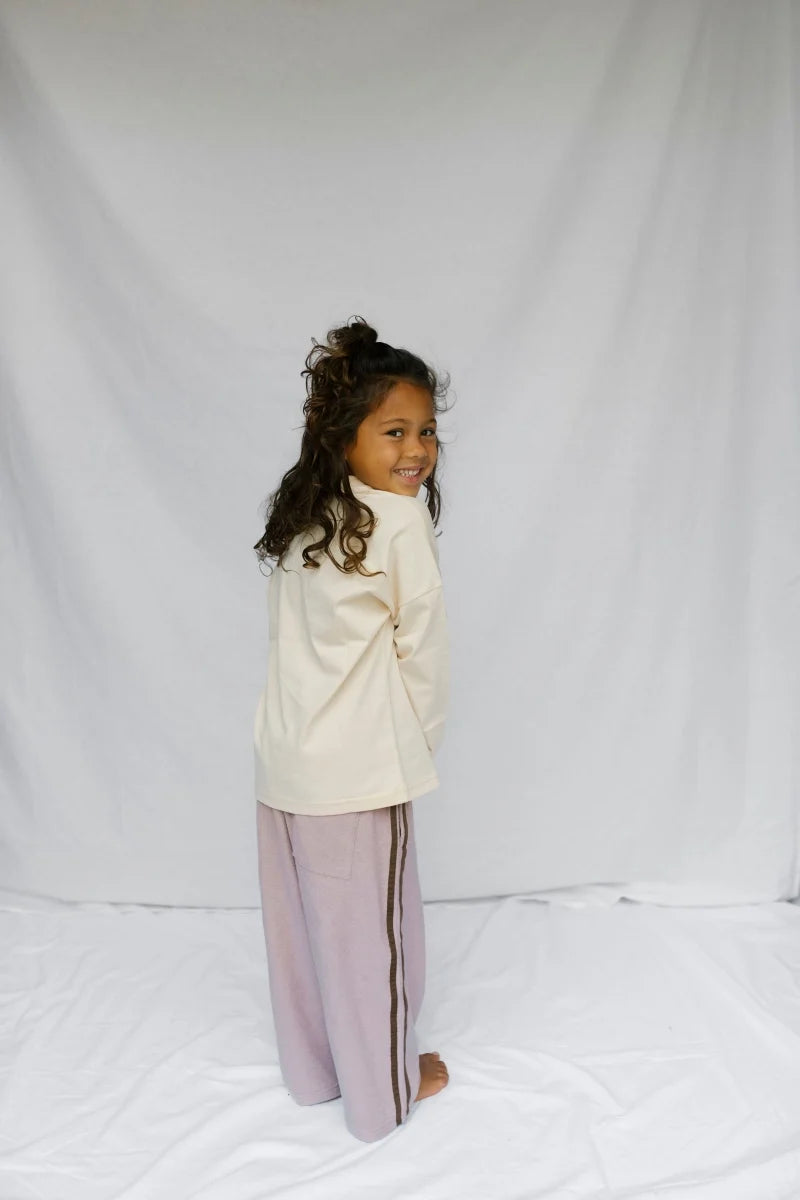 A young girl with curly brown hair stands in profile against a white fabric backdrop, wearing a cream long-sleeve top and light lavender wide-leg pants with dark side stripes.