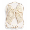 A large cream-colored cotton bow with tiny gold polka dots, presented on a decorative white and gold display card. The brand name 'cléra' is visible at the bottom of the card.