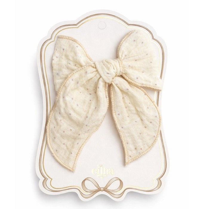 A large cream-colored cotton bow with tiny gold polka dots, presented on a decorative white and gold display card. The brand name 'cléra' is visible at the bottom of the card.