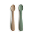 Two silicone spoons, one beige and one green, on a white background with 'mushie' branding.