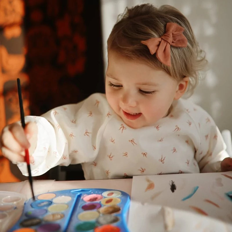 Child painting with a watercolor set, wearing a white shirt with bows patterns and a pink bow in hair.