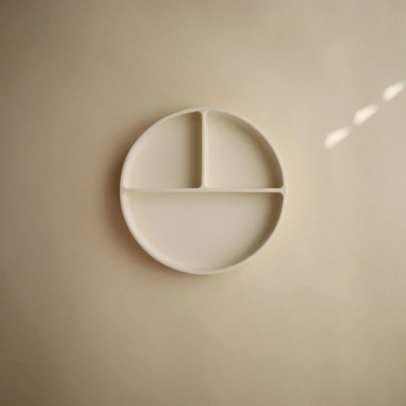 A minimalist off-white circular wall-mounted plate with three divided sections, shown against a matching beige wall with soft natural light shadows.