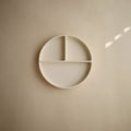 A minimalist off-white circular wall-mounted plate with three divided sections, shown against a matching beige wall with soft natural light shadows.