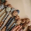 A close-up flat lay of multiple pacifier clips and beaded straps in soft pastel tones, including blue, gray, pink, beige, and brown wood. The clips have silver metal hardware and the beads are smooth and matte.