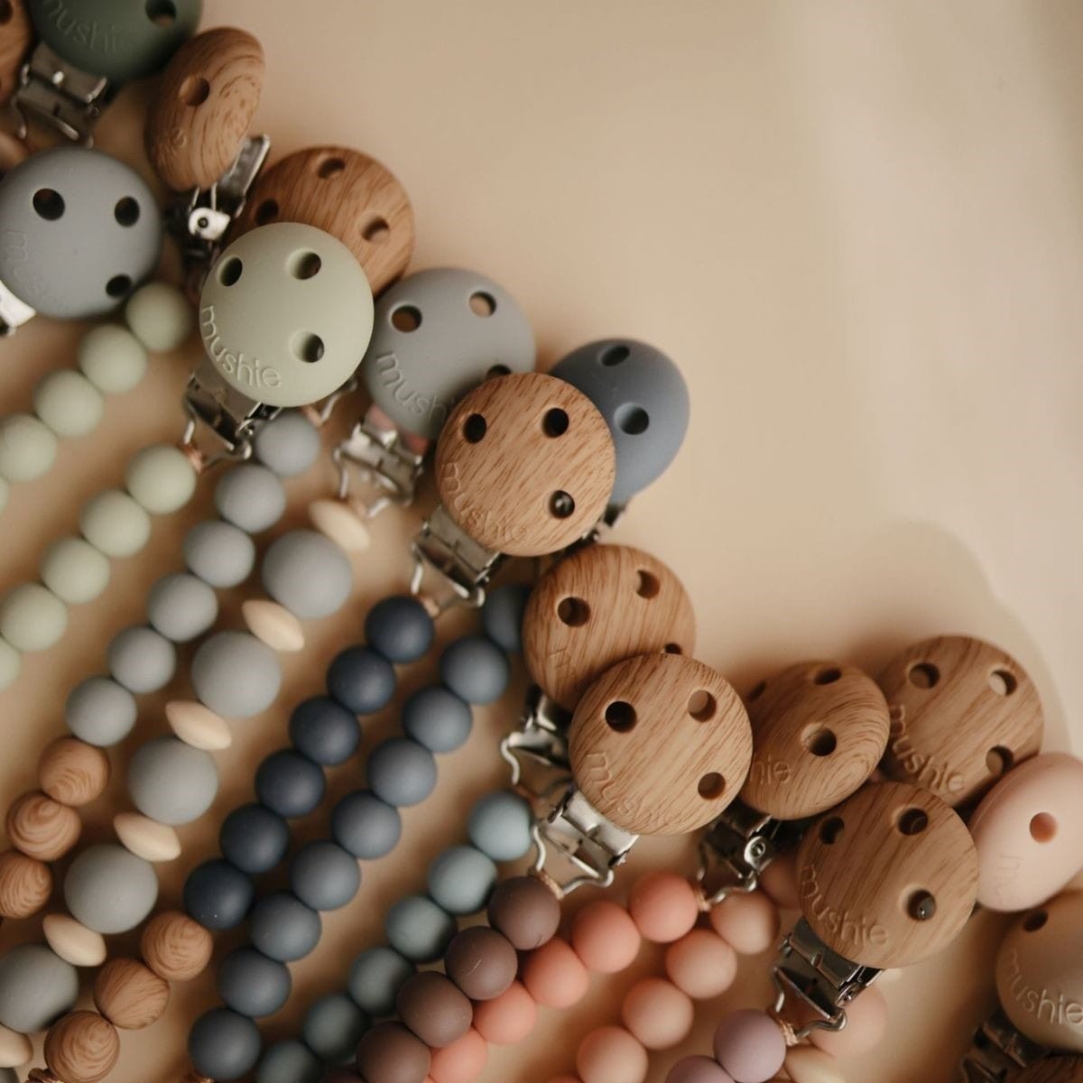 A close-up flat lay of multiple pacifier clips and beaded straps in soft pastel tones, including blue, gray, pink, beige, and brown wood. The clips have silver metal hardware and the beads are smooth and matte.