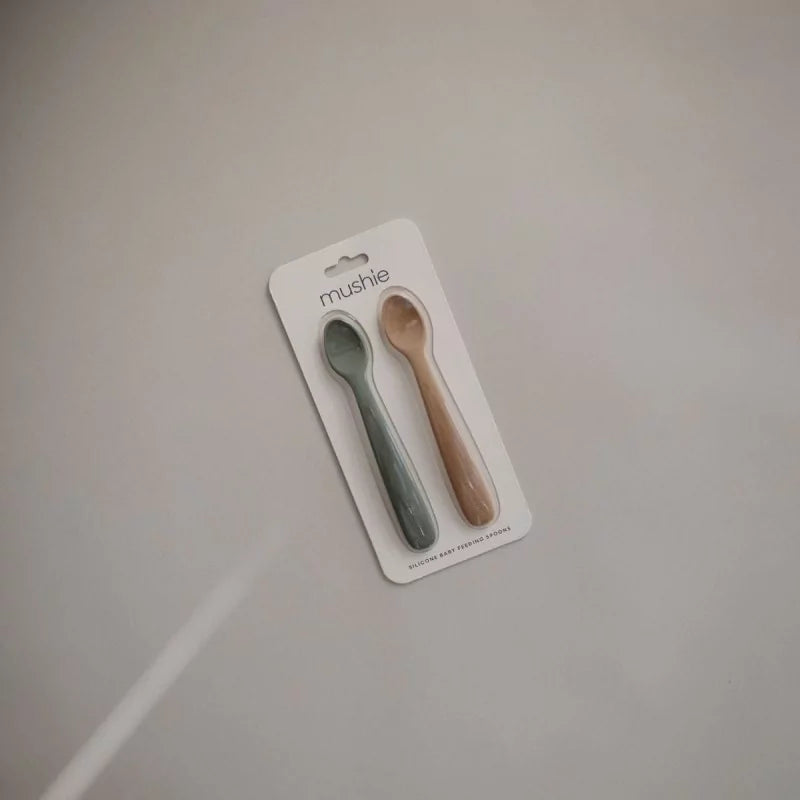 Two baby spoons in packaging labeled 'mushie' on a plain background