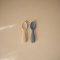 Two baby spoons, one beige and one gray, on a beige background