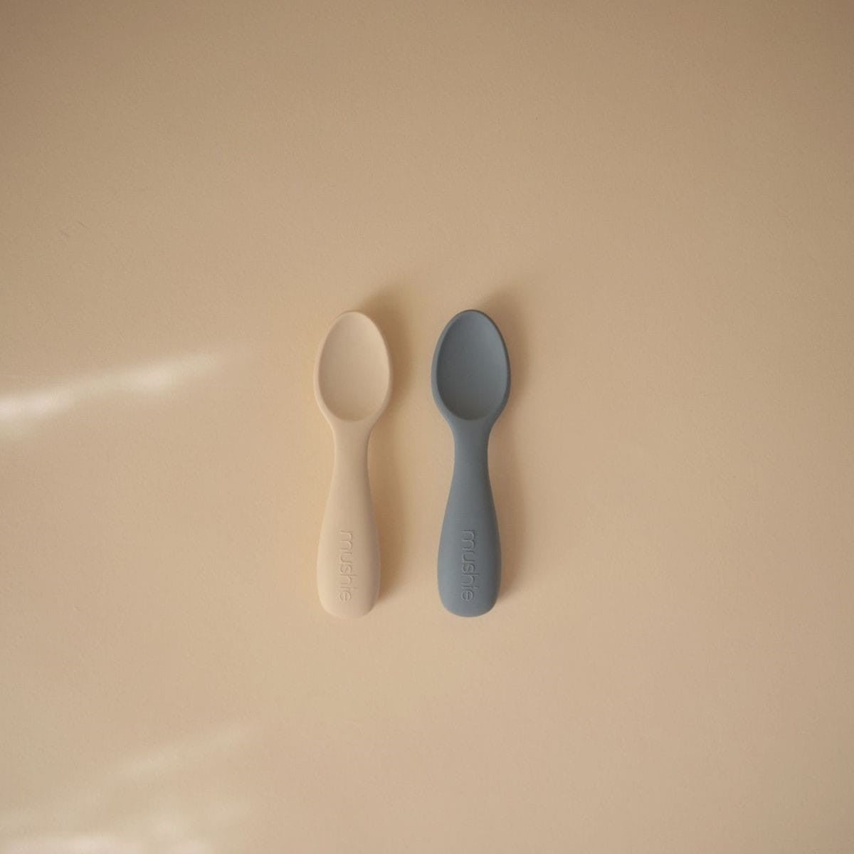 Two baby spoons, one beige and one gray, on a beige background