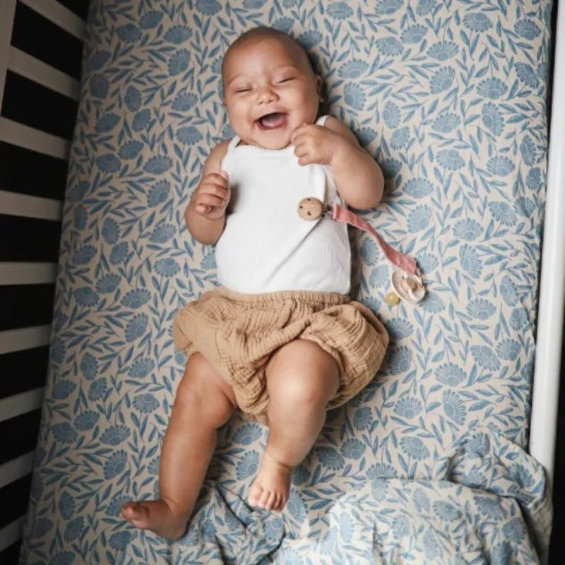 Baby lying on a mushie blue floral blanket with a white outfit and beige shorts