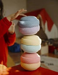 A stack of four pastel macaron-shaped plush pillows in blue, pink, yellow, and orange, with a person in a red sweater touching the top one, set on a red table in a warm indoor environment.
